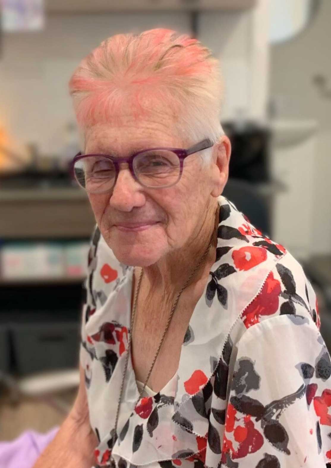 An elderly woman with pink hair sitting in a chair.